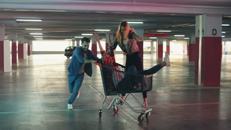 young cheerful caucasian man having fun with two girls while carrying them in the trolley in a empty parking