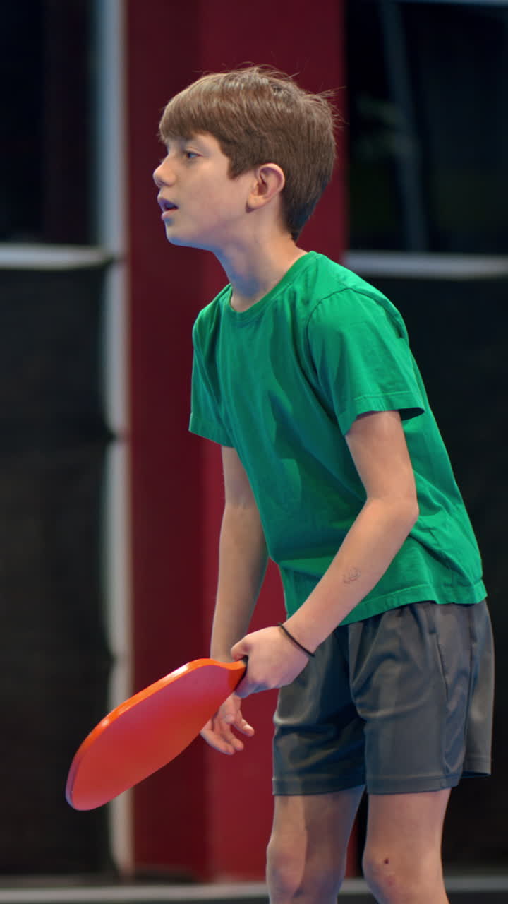 A boy playing pickleball on a blue, inside court. Vertical