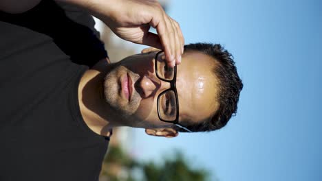 Man-standing-outdoors-on-a-sunny-day,-putting-on-his-sunglasses-with-one-hand,-and-a-blurred-background-of-trees-and-buildings-behind-him