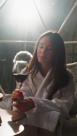 woman with glass of wine sits on balcony. relaxed lady in soft bathrobe enjoys date with boyfriend sitting in cozy armchair at glamping house at night