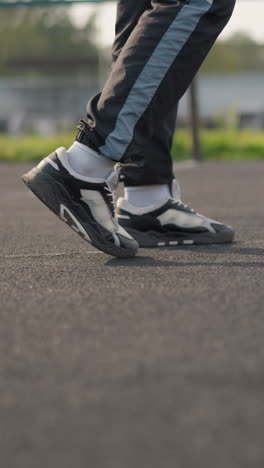 leg view of person wearing joggers and sneakers walking on court with blurred background featuring bar fence and greenery, focus on footwear and court surface in outdoor environment
