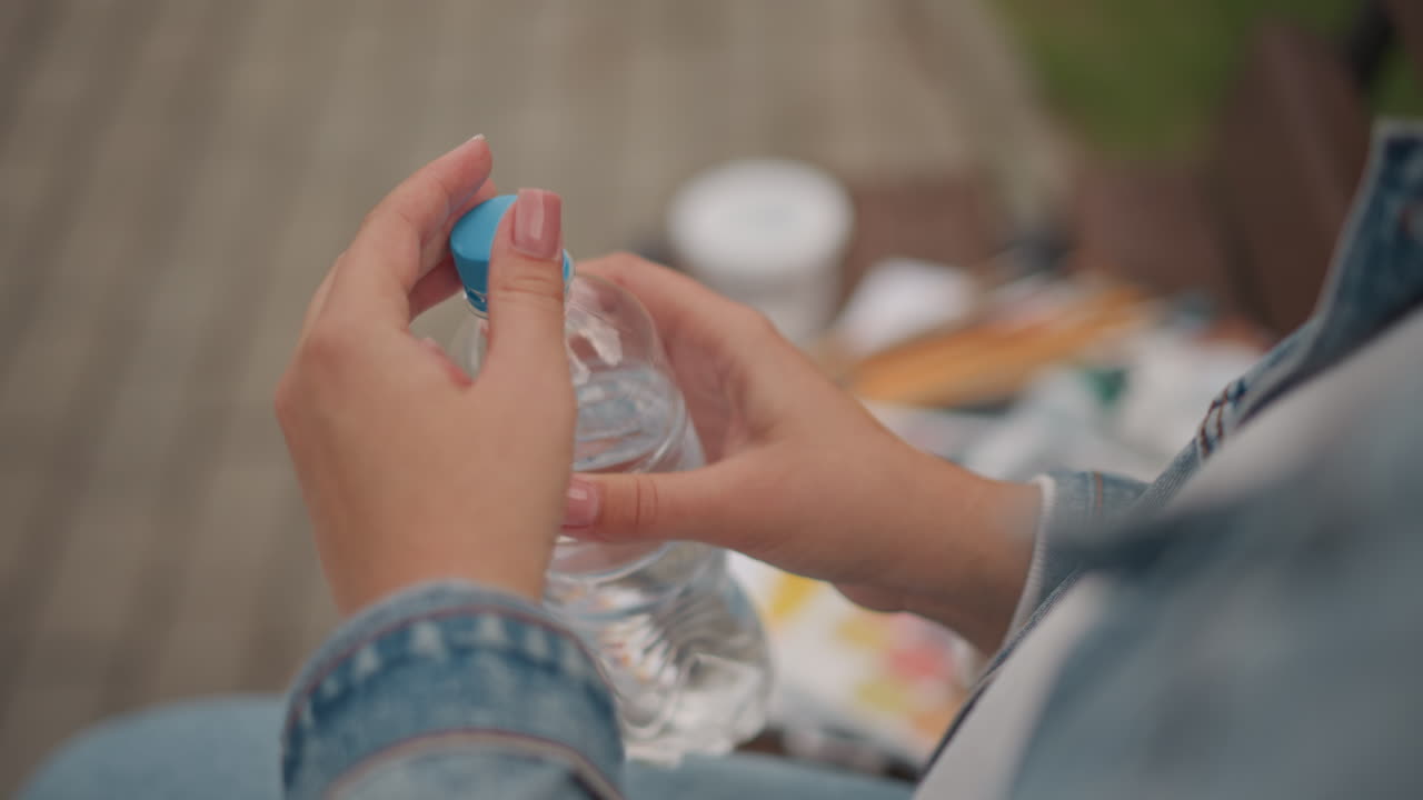 Donna bianca che apre una bottiglia d'acqua di plastica, primo piano delle mani che avvitano il tappo blu, maniche di jeans e unghie curate, oggetti da picnic sfocati su una panca, momento di idratazione spontaneo, luce solare estiva