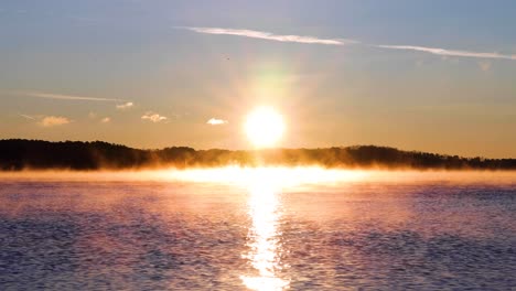 4k medium close up sunrise over a lake with mountain in the background, sun-ray sprinkling through water, blue and orange sky, fog on water