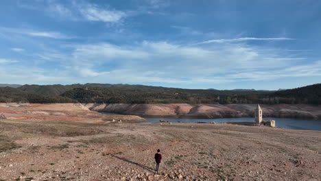 vista aérea que se eleva sobre el hombre de pie en la tierra árida de la sequía mirando a través de la iglesia de sant roma en el embalse de panta de sau