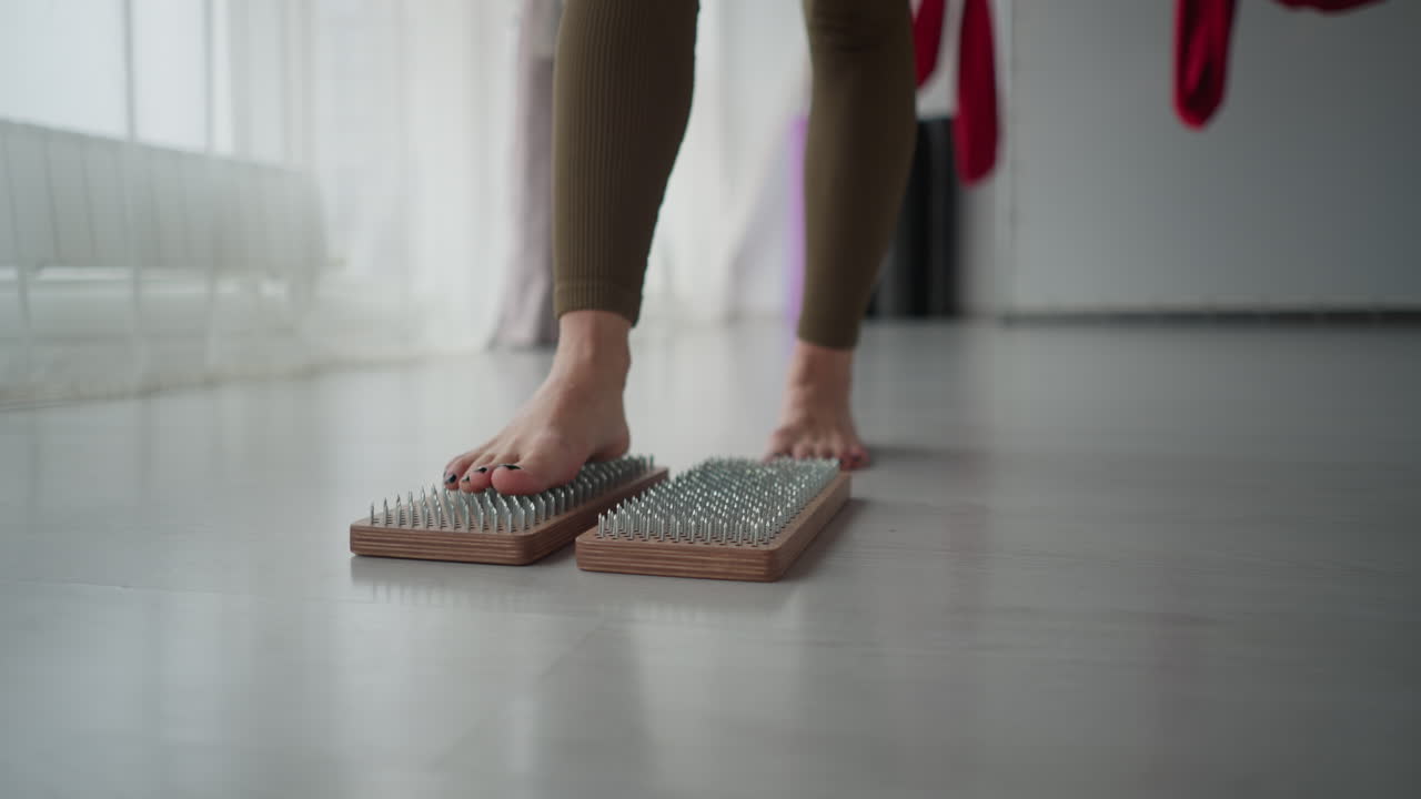 Fitness coach balances bare feet on wooden nail pad blocks barefoot on yoga mat in bright studio surrounded by red aerial silks and floor to ceiling windows during mindful wellness session