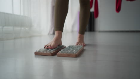 fitness coach balances bare feet on wooden nail pad blocks barefoot on yoga mat in bright studio surrounded by red aerial silks and floor to ceiling windows during mindful wellness session