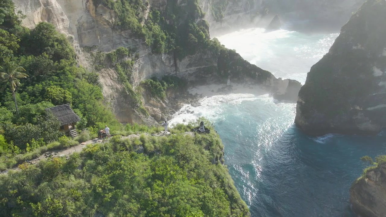 Tomada aérea cinematográfica da icônica costa da Praia do Diamante em Nusa Penida, Bali, apresentando águas turquesa, falésias de calcário e vegetação exuberante durante uma manhã ensolarada.