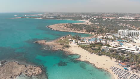 Aguas-Turquesas-Claras-Y-Poco-Profundas-Que-Complementan-La-Prístina-Costa-De-La-Playa-De-Nissi,-En-Ayia-Napa,-Chipre---Toma-Panorámica-Aérea-Alta-Y-Lenta