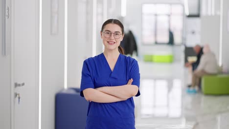 retrato de una chica morena segura y feliz con gafas en un uniforme azul el médico cruza los brazos sobre su pecho y posa en el corredor de una clínica de luz moderna