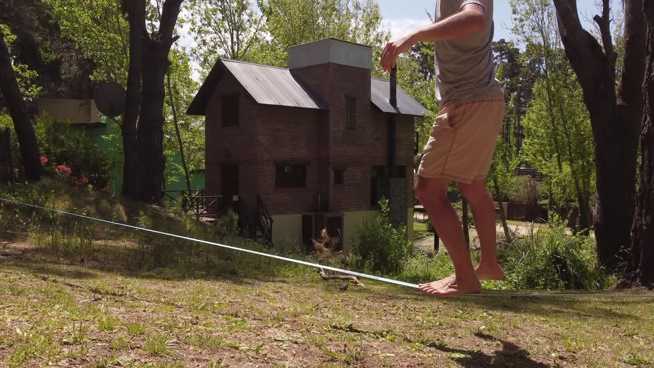 Close up shot of man balancing on slack line in forest with house in background during summer