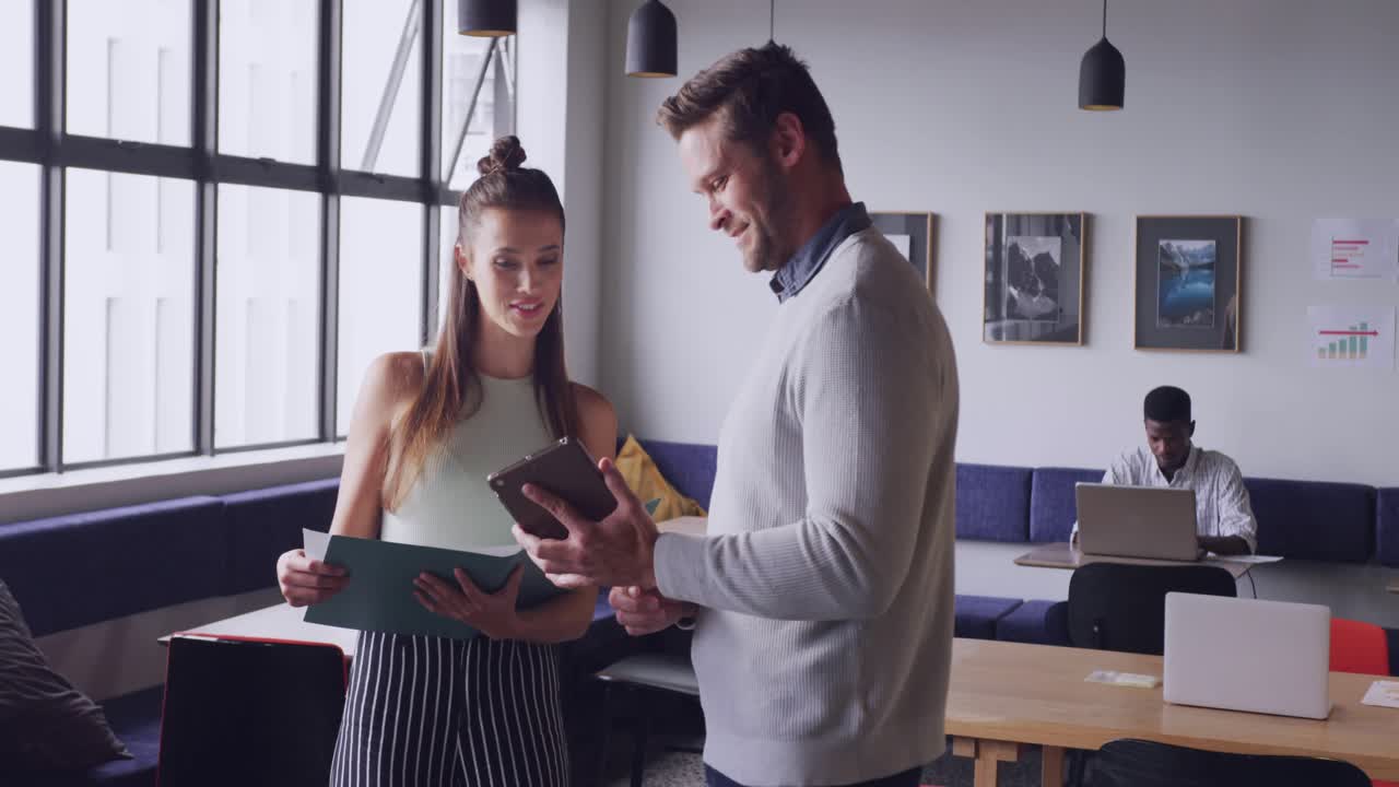 Male tapping tablet forming HUD, female comparing folder in coworking for business review