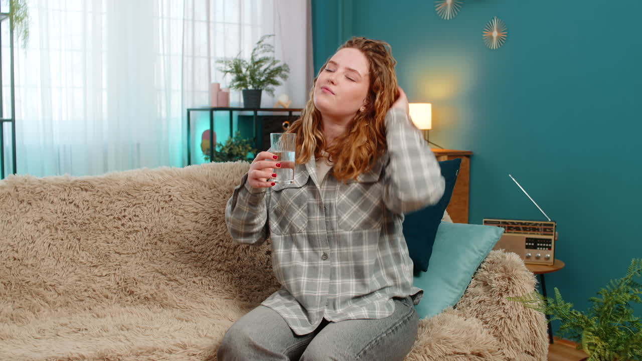 Woman sitting on sofa drinking water feeling refreshed after workout or hot summer day relaxation