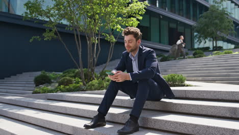 handsome manager typing message on smartphone sitting stairs outdoors zoom out.