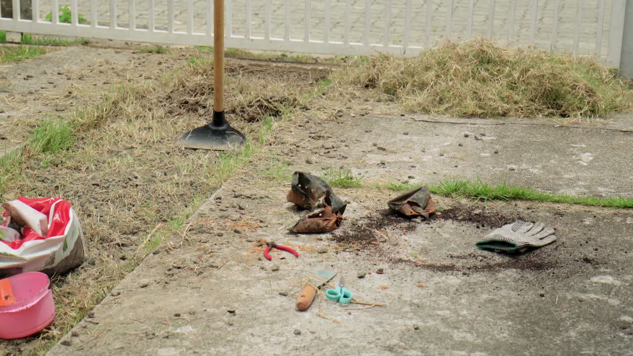 Scene Take of a camera tilting up on dirty gardening equipment, including a hoe, and a pile of pulled dry weeds resting on the ground next to a white fence after backyard maintenance