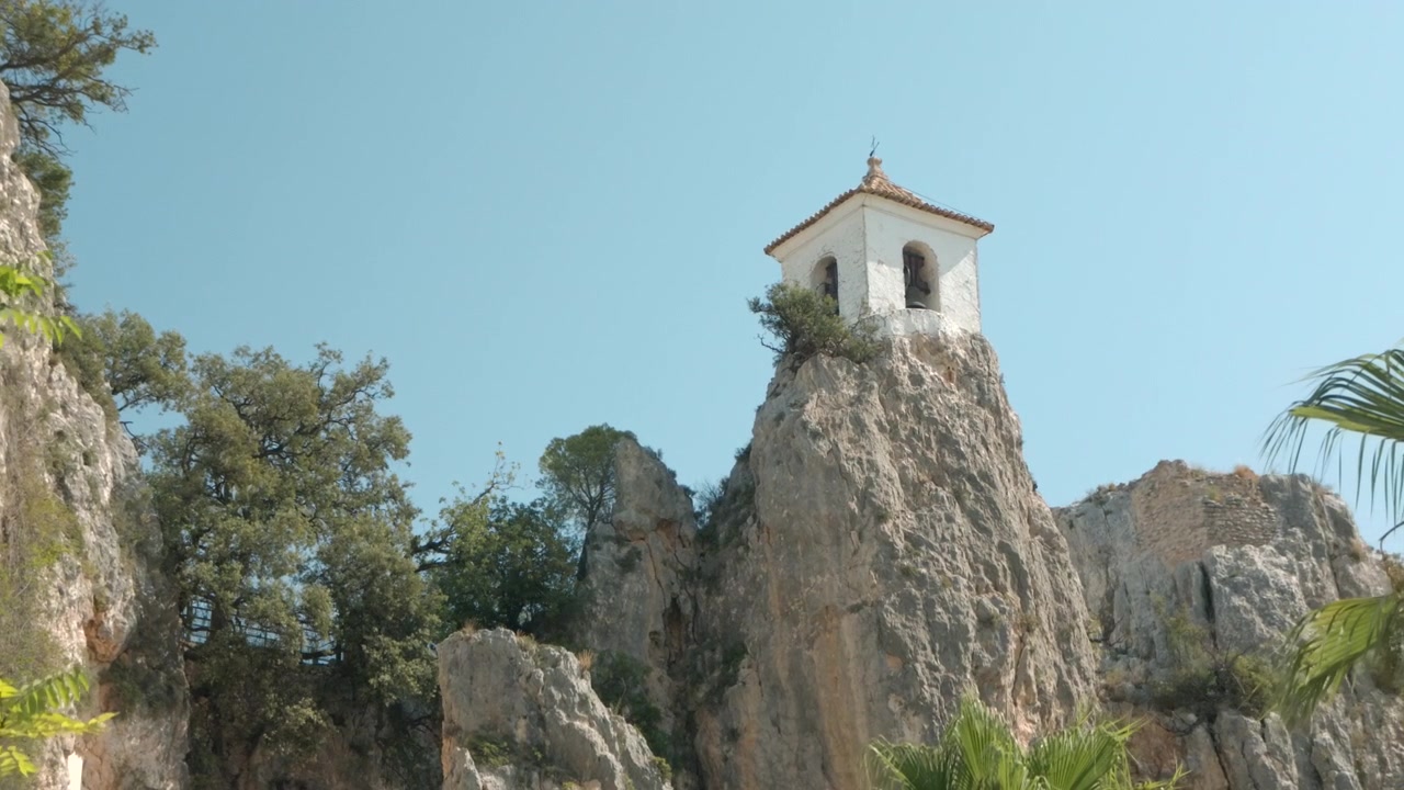 Paesaggio roccioso luminoso con piccola cappella bianca in cima a un'alta guglia di pietra, circondata da scogliere aspre, alberi sparsi, foglie di palma e cielo azzurro limpido, creando un'ambientazione esterna scenografica e calma.