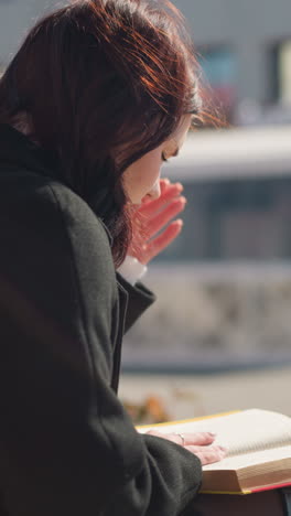 야외에서 책을 읽는 데 집중하는 여자의 근접 후면 뷰, 그녀의 머리카락이 부드럽게 바람에 흔들리면서, 그녀는 빛이 부드러운 도시 환경에서 장면을 강조하는 동안 다음 페이지를 뒤집습니다.