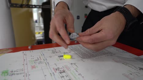 Man-in-uniform-taking-earplugs-out-from-plastic-wrapping-and-laying-them-on-table-with-ships-engine-room-drawings---Closeup-of-hand-and-earplugs