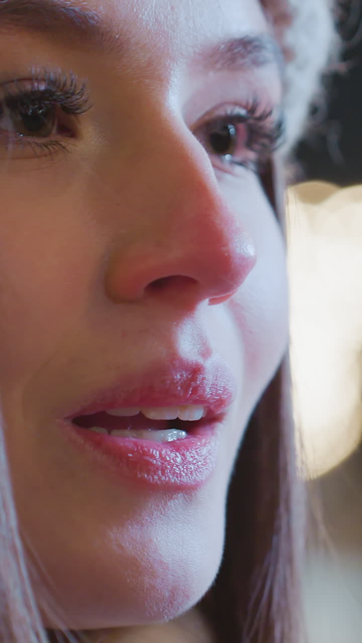 Close-up of young woman with hands near mouth, exhaling warm breath in cold winter air, visible breath vapor highlights freezing atmosphere, background features soft bokeh lights