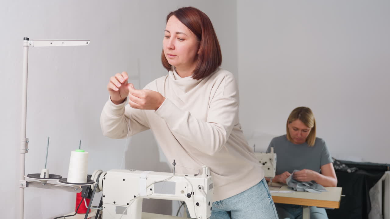 Woman seamstress carefully fixing white thread on electric sewing machine in bright workshop while colleague works on clothing in background, highlighting expertise, precision, and focus