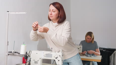 woman seamstress carefully fixing white thread on electric sewing machine in bright workshop while colleague works on clothing in background, highlighting expertise, precision, and focus