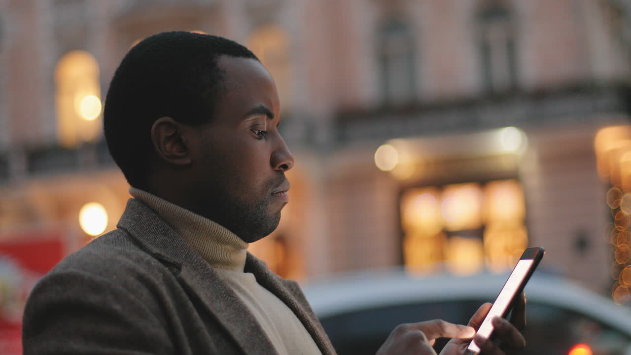 Side view of African American young man texting on the smartphone in the street in the evening