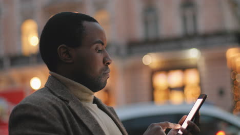 Side-view-of-African-American-young-man-texting-on-the-smartphone-in-the-street-in-the-evening