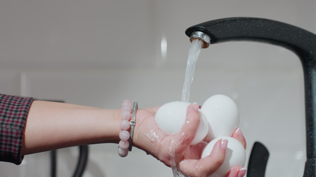 primer plano de una mujer con la mano en ropa a cuadros lavando tres huevos blancos bajo agua corriente, lleva una pulsera de cuentas mientras enjuaga los huevos en un fregadero de cocina moderno