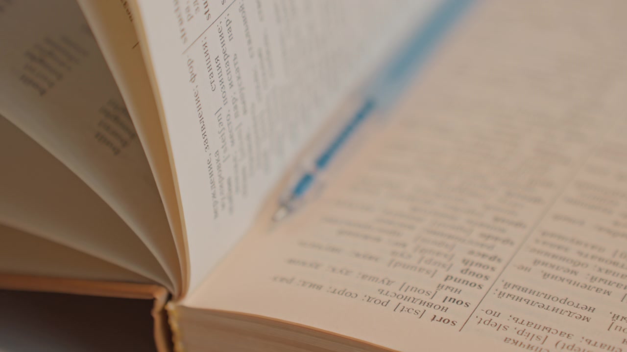 Close up of open dictionary with bilingual text. Camera slowly zooms in and pans across detailed word entries. Soft lighting emphasizes paper texture and text clarity