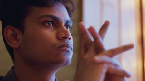 Close-up of young man in profile resting chin on clasped hands, looking out with serious thoughtful gaze, soft light illuminating his face, suggesting deep reflection, calm emotion, and quiet intensity