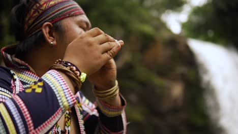 orbit-shot-of-an-Indigenous-person-playing-Kubing-tribal-instrument,-behind-is-a-waterfall