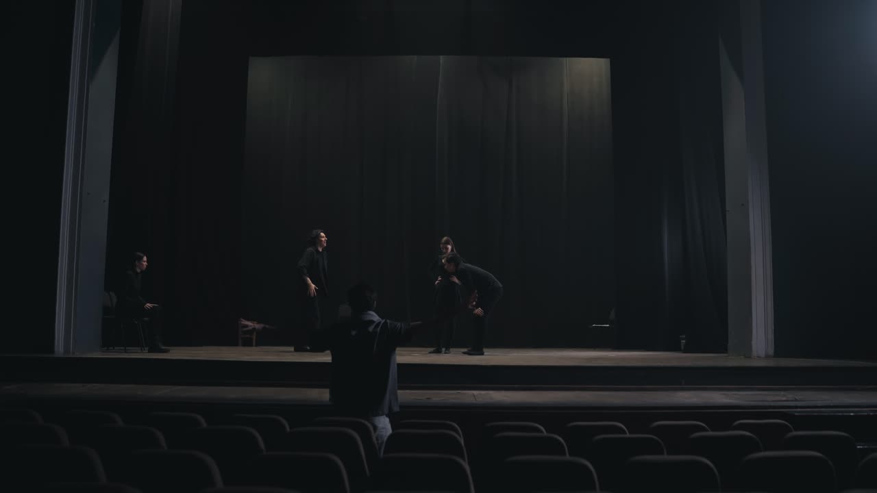 задний вид молодого мужского продюсера, стоящего в зале и показывающего, как перемещать актеров и что делать на сцене во время репетиции в пустом театре с черными занавесками