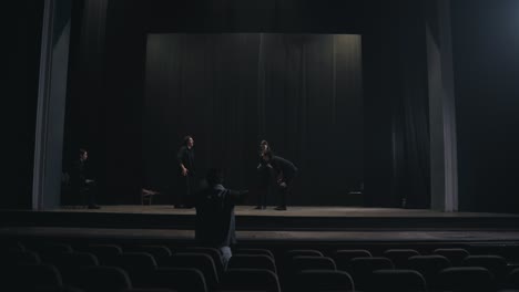 rear view of a young male production director standing in the hall and showing how to move the actors and what to do on stage during a rehearsal in an empty theater with black curtains