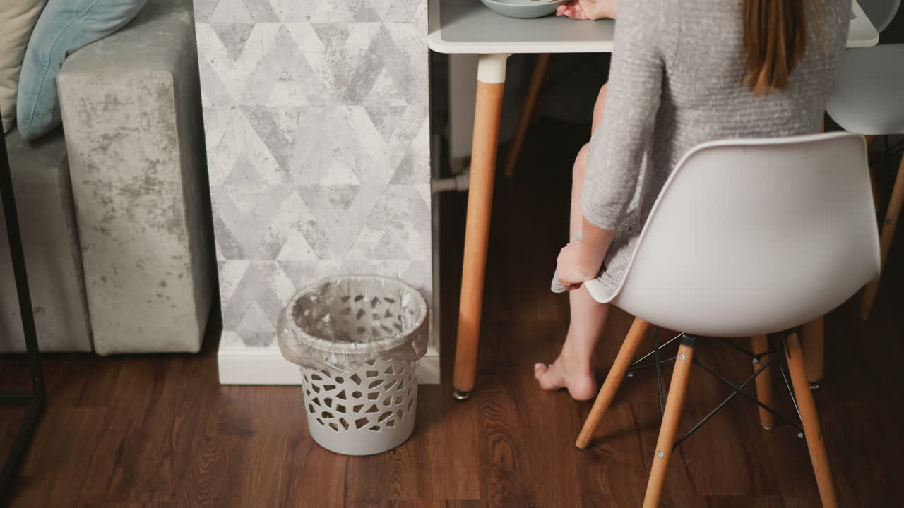 Back view of lady discarding potato peels into trash can while seated at dining table, plastic-lined bin placed against patterned wall, cozy home interior with wooden floor and cushioned sofa nearby