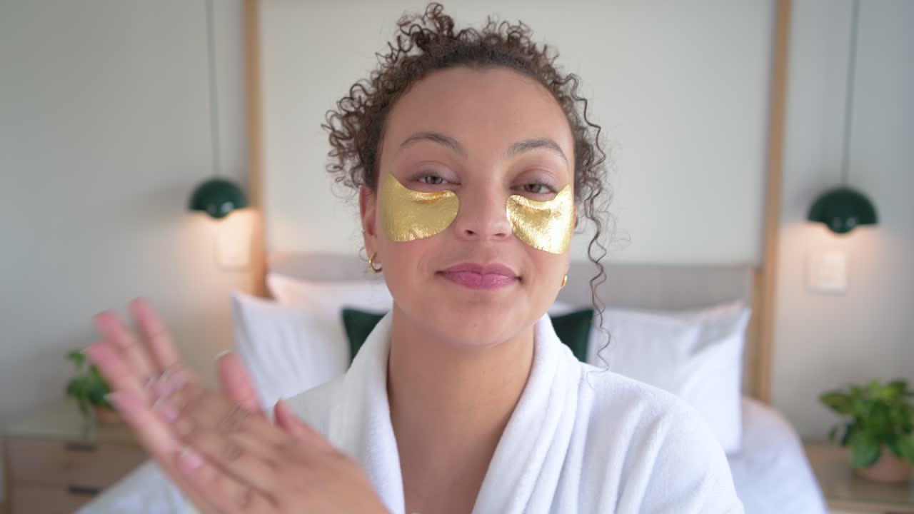 Adult woman in white bathrobe placing and pressing gold under-eye patches on bed for skincare