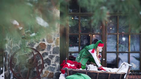 Two elves in green costumes sitting in wooden sleigh outdoors. Happy elves playing snowballs together on house background in winter