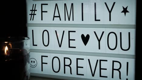 a lit glass candle in front of a billboard with the text family love you forever, a hashtag and some symbols