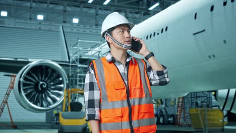 ingeniero masculino asiático con casco de seguridad hablando en teléfono inteligente mientras está de pie con el avión en el hangar