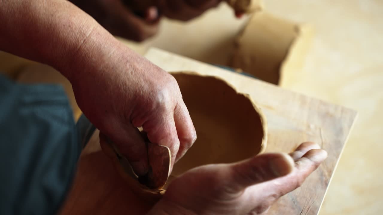 Seen from over the shoulder, skilled hands carefully shape a clay bowl on a wooden board during a pottery lesson in natural light