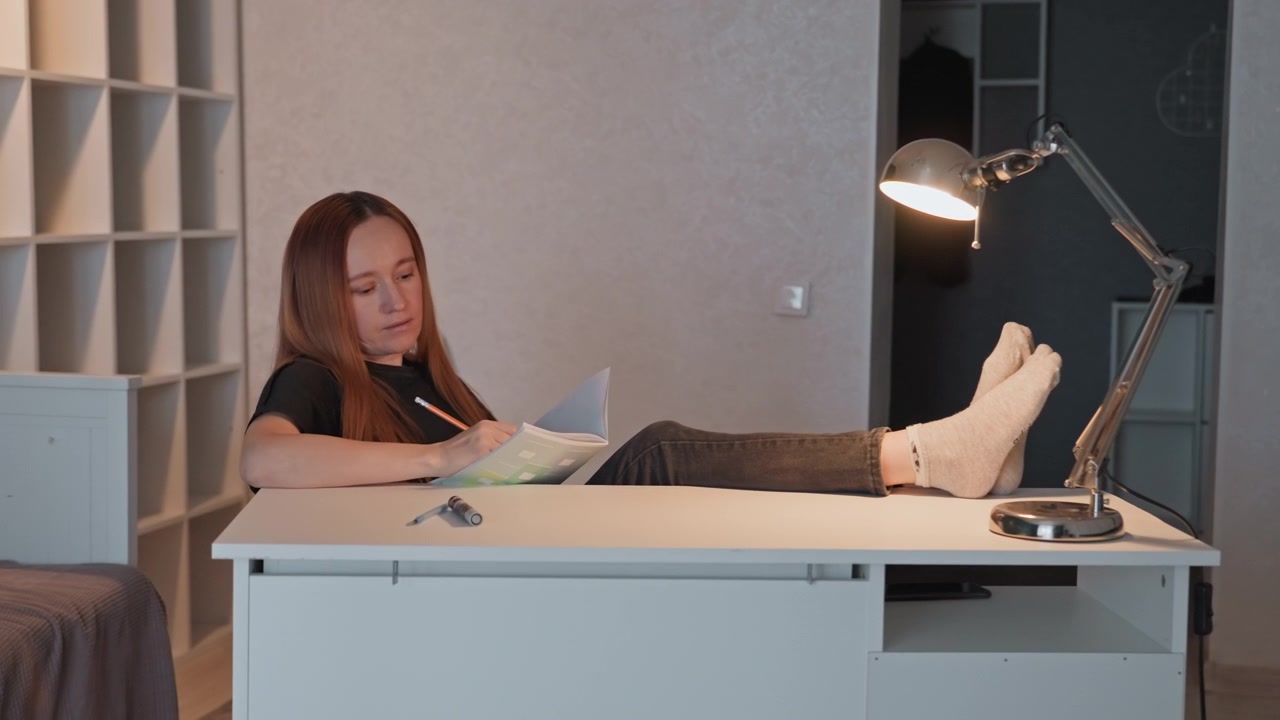 Young woman lies on white desk reading and writing in open book. Desk lamp illuminates her workspace feet rest casually at edge of table. Shelving units visible behind subject