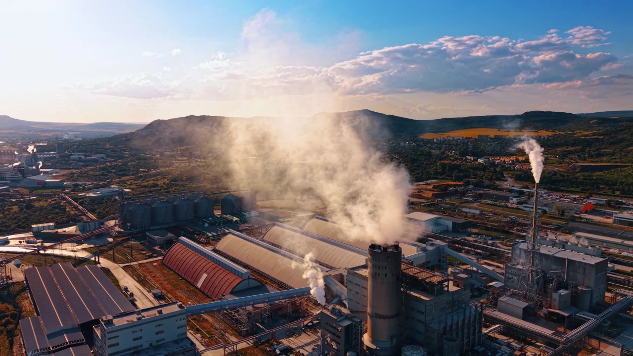 Nuvole di vapore che si innalzano sopra un impianto industriale vicino a colline boscose. Una vista elevata mostra ciminiere, capannoni di produzione, silos di stoccaggio e infrastrutture ferroviarie.