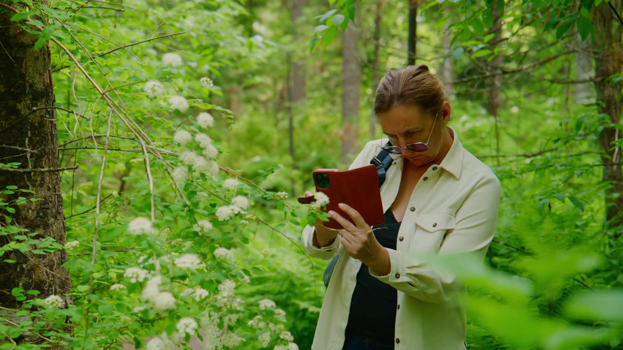 Donna cammina attraverso una lussureggiante foresta verde, tenendo un taccuino rosso. Esamina un fiore bianco con curiosità e attenzione. La luce del sole filtra tra gli alberi, creando un'atmosfera pacifica e naturale