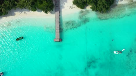 Maldives,-beautiful-peaceful-white-sand-beach-with-wooden-deck-,tropical-trees-and-boats-floating-in-the-calm-turquoise-water