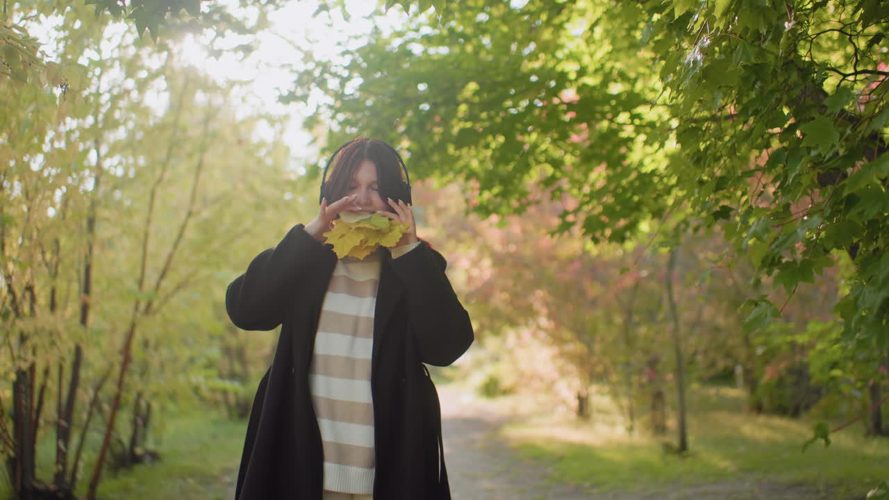 Medium view of young girl biting yellow leaves while preparing to put on headset during autumn walk in bright forest pathway, playful expression with sunlight filtering through green foliage