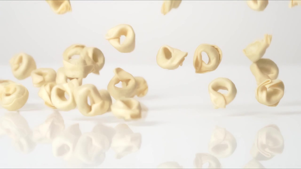 Tortellini freschi non cotti che rimbalzano su un tavolo bianco in slow motion, in primo piano macro, prima di posarsi.