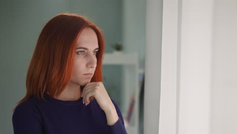 pensive redhead woman looks out room window touching chin