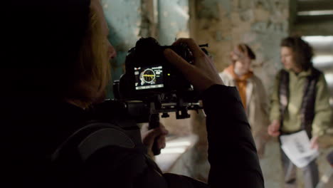 Rear-View-Of-Cameraman-Recording-His-Coworkers-Reading-A-Script-In-A-Ruined-Building-1