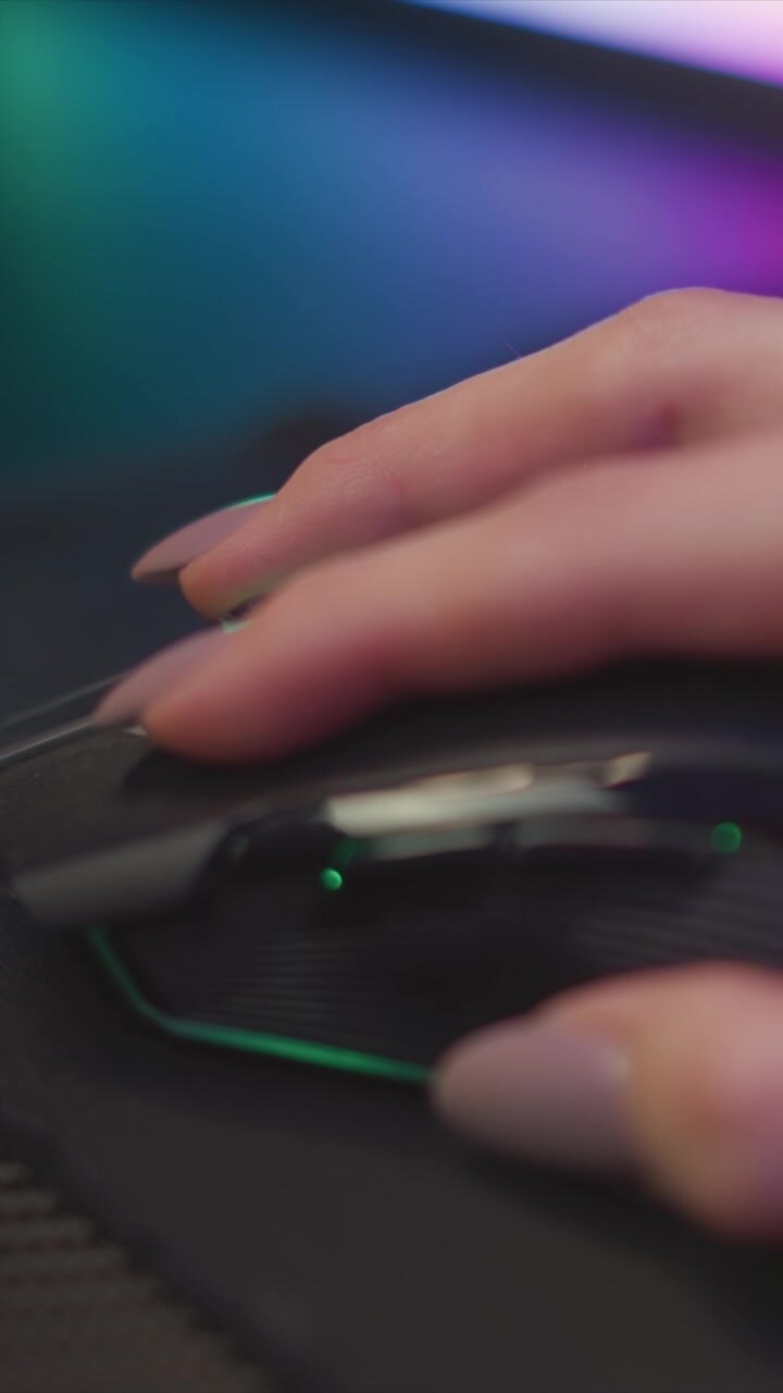 Hand with manicured nails moves gaming mouse across desk focusing on interaction between fingers and device. background features soft gradient lighting in green and purple hues creating