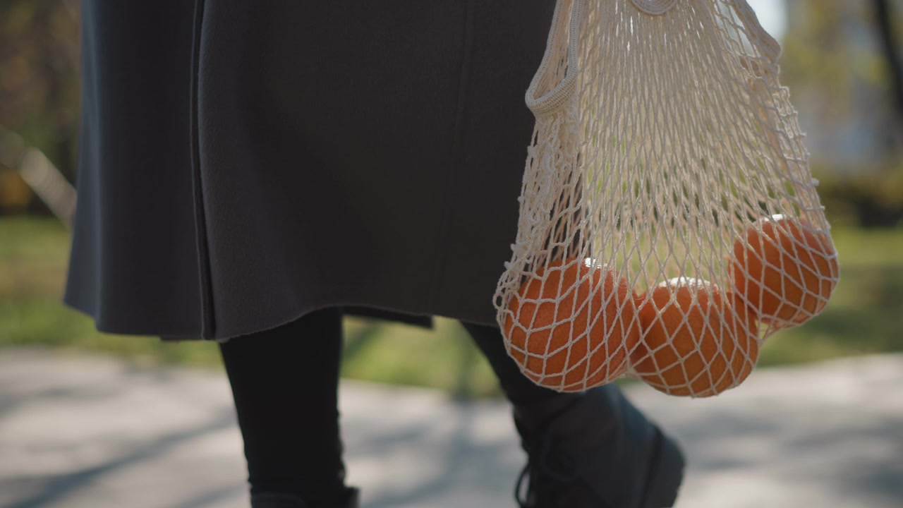 Primo piano di una persona che cammina con diverse arance in una borsa a rete, indossando abiti scuri. Il soggetto porta una borsa a rete per la spesa contenente frutti rotondi, su uno sfondo di parco all'aperto con vegetazione.