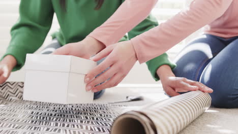 Low section of biracial mother and adult daughter wrapping christmas gift at home, slow motion