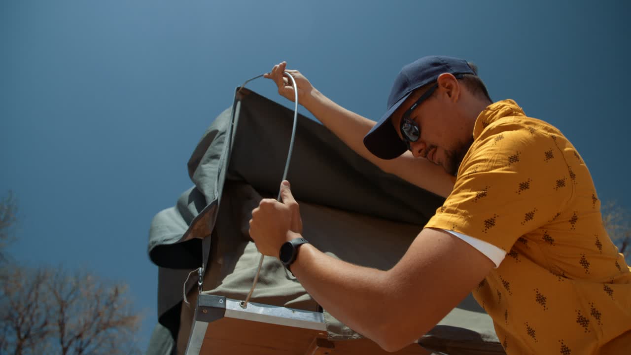 Hand held tracking shot of a off-roading Caucasian male tourist in Africa as he walk towards and climbs up ta ladder of a off-roading vehicle and proceeds to insert a rooftop canvas tent door peg.